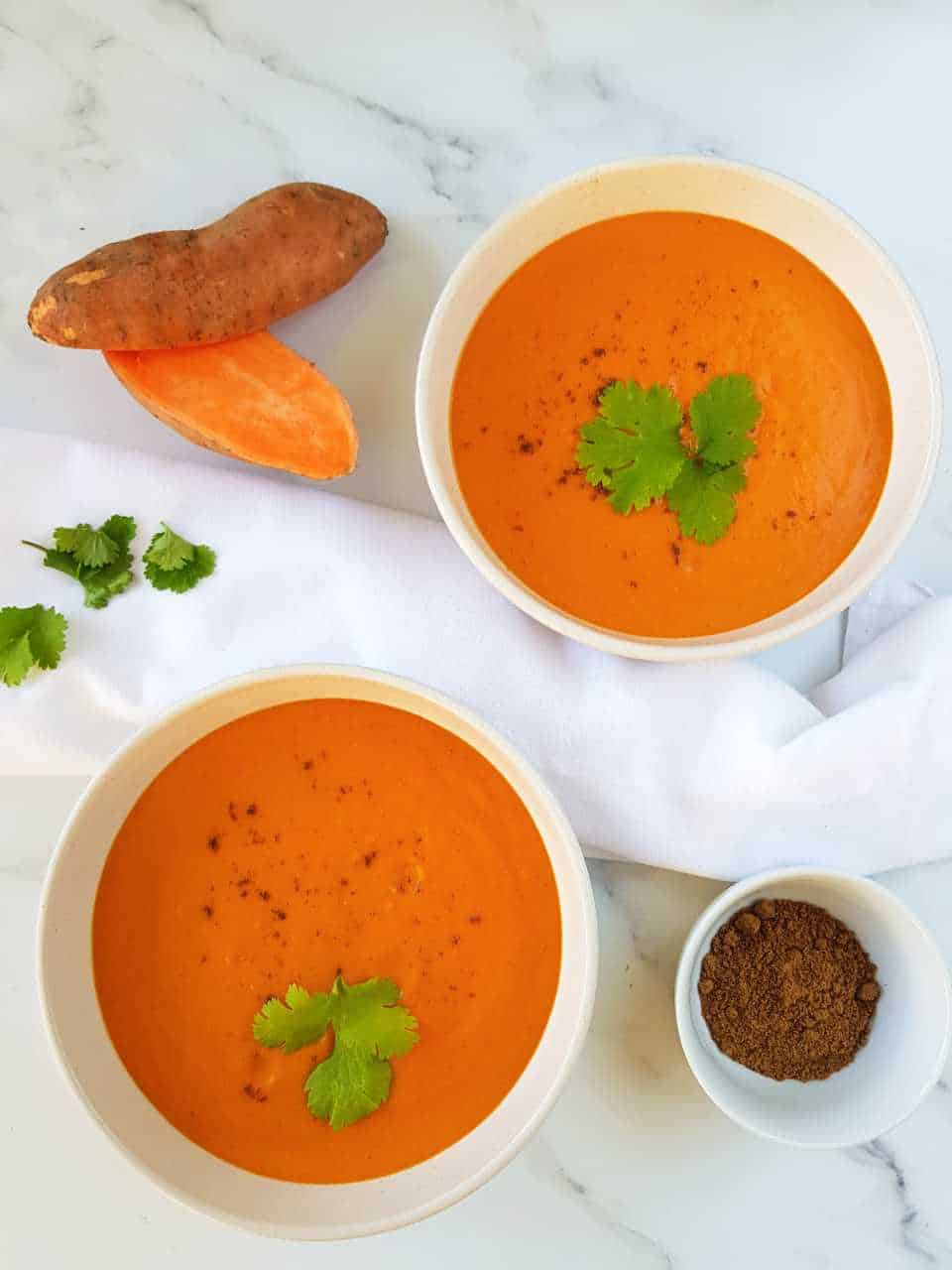 Sweet potato and red pepper soup in white plates on a marble table with sweet potatoes and a bowl of spices on the side.