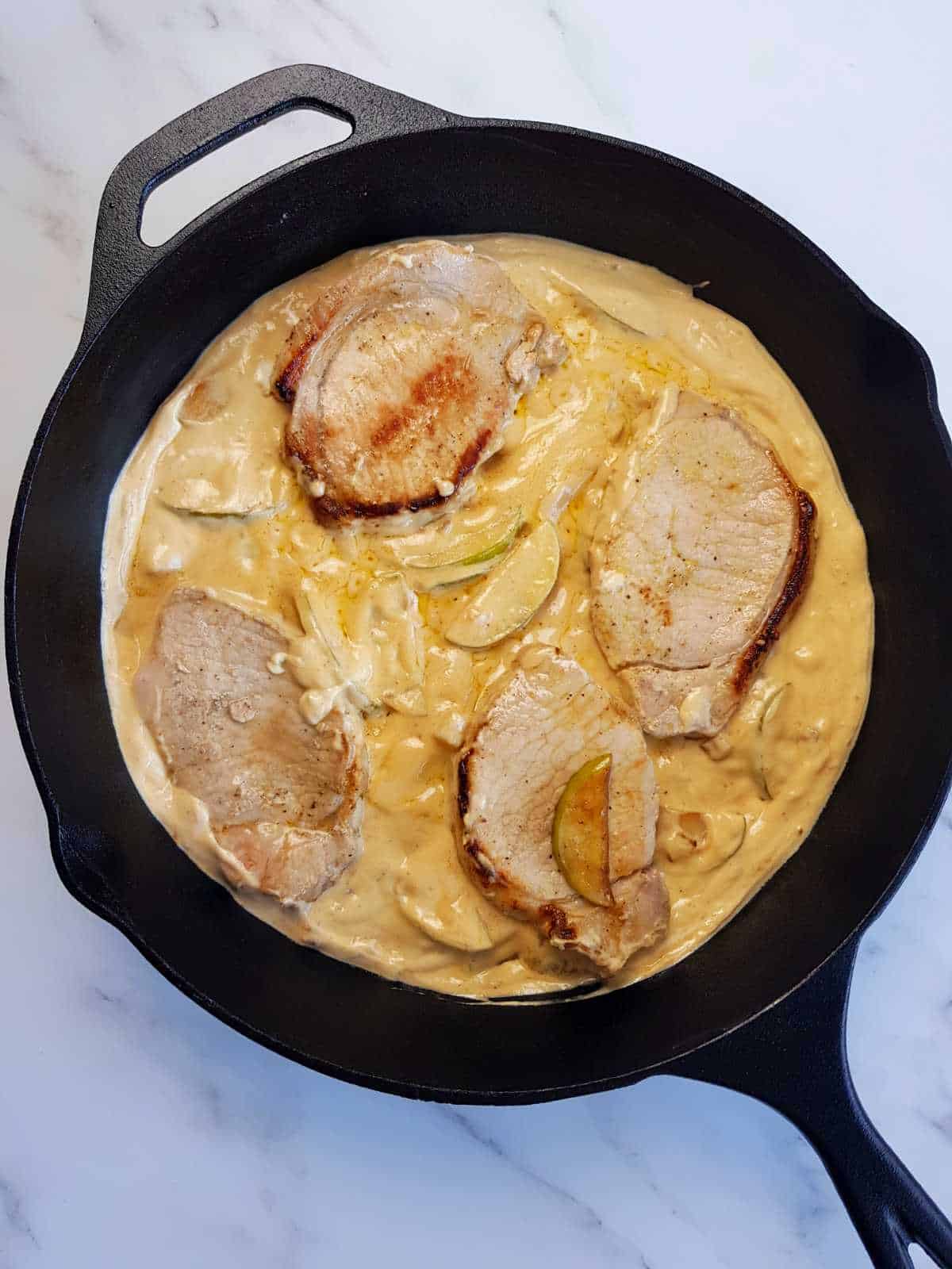 Pork chops in apple cream sauce in a cast iron skillet.